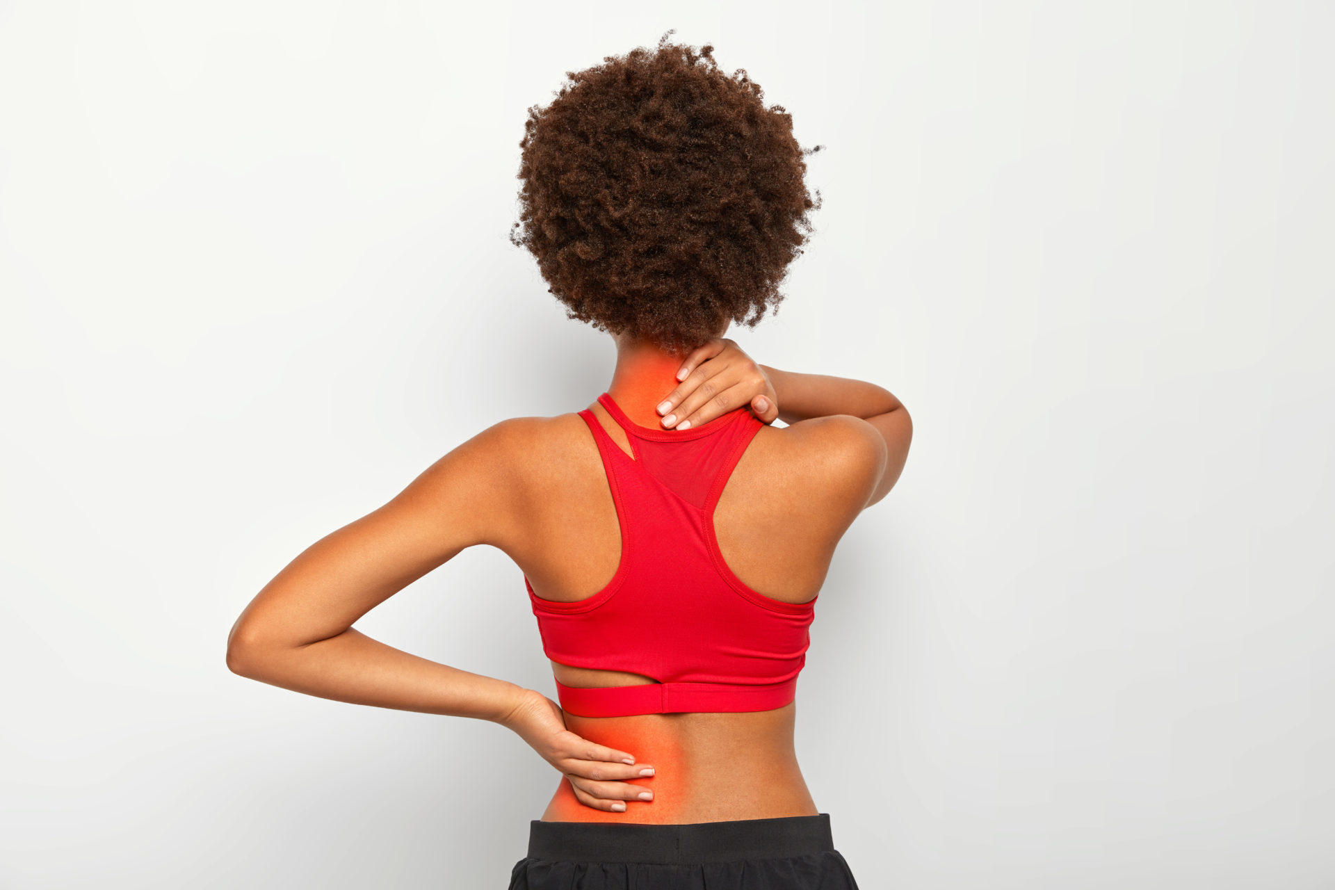 Brunette dark skinned female model has joint pain in back and neck, has health problem, dressed in active wear isolated over white studio wall. Medical and osteopathy concept. Spine disease or illness