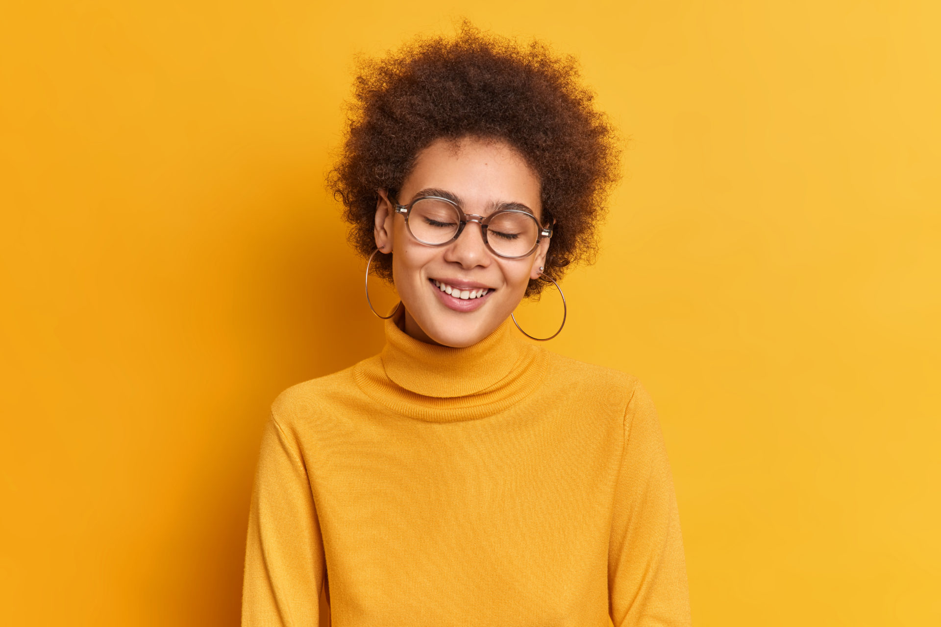 Horizontal shot of happy sincere millennial girl with curly hair natural beauty smiles gently with closed eyes gets compliment enjoys life wears casual poloneck isolated over yellow background Woman happy smiling